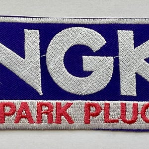 May include: Embroidered patch with the text "NGK SPARK PLUGS." The patch features white embroidered lettering on a blue background, with red embroidered "SPARK PLUGS" at the bottom. The patch has a white border.