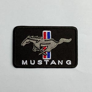 Peut inclure: Écusson rectangulaire noir avec le logo Mustang : un cheval gris au galop et une bande verticale rouge, blanche et bleue. Le mot "MUSTANG" est brodé en blanc en bas.