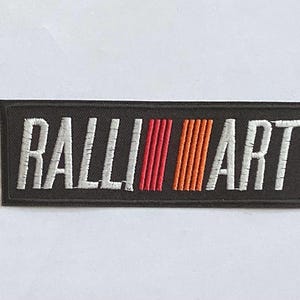 Puede incluir: Parche rectangular negro con el texto bordado "RALLI ART" en blanco. El diseño incluye tres rayas verticales en rojo y naranja. Ideal para entusiastas de los coches o ropa de automoción.