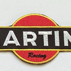 以下が含まれることがあります： 白い「MARTINI」の文字が刺繍されたパッチ。黒い長方形の背景に金色の縁取りが施されています。パッチは赤い円形の背景と赤い「Racing」の文字が特徴です。