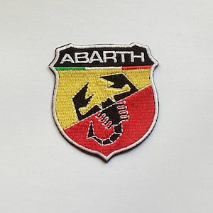 May include: Embroidered patch featuring the Abarth logo. The patch is shield-shaped with a black border and the word "ABARTH" at the top. The design includes a yellow and red background with a black scorpion in the center.