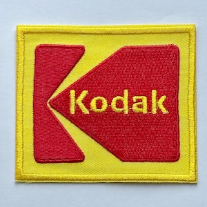 Puede incluir: Parche bordado con el logotipo de Kodak. El parche tiene un borde y fondo amarillos, con una forma de "K" roja estilizada y la palabra "Kodak" en amarillo. El parche es cuadrado.