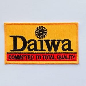 May include: Embroidered patch with the "Daiwa" logo in black on a yellow background. The patch has a red border at the bottom with the text "COMMITTED TO TOTAL QUALITY". A black and white swirl design is above the "Daiwa" text.