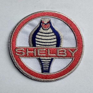 Op de afbeelding: Ronde geborduurde patch met een cobra-embleem in blauw, wit en grijs, omcirkeld door een rode ring. Het woord "SHELBY" is rood geborduurd. De patch heeft een getextureerd oppervlak en is bedoeld voor textiel.