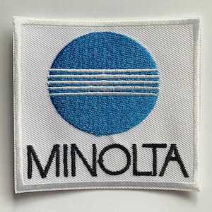 Pode incluir: Patch quadrado branco com um círculo azul e três linhas horizontais brancas. A palavra "MINOLTA" é bordada em preto abaixo do círculo. O patch tem uma borda branca.