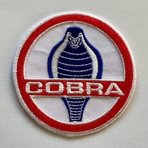 Peut inclure: Écusson brodé rond avec un motif de cobra bleu et blanc sur fond blanc. L'écusson est bordé d'un anneau rouge, avec le mot "COBRA" en lettres blanches.