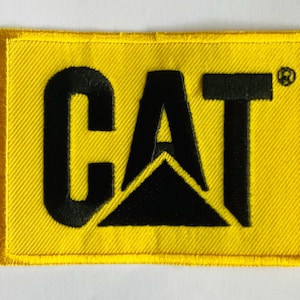 Könnte beinhalten: Ein gelber rechteckiger Aufnäher mit dem schwarzen "CAT"-Logo. Das Logo enthält das Wort "CAT" in fetten, schwarzen Buchstaben, mit einem schwarzen Dreieck unter dem "A". Der Aufnäher hat einen gelben Rand und ein strukturiertes Aussehen.