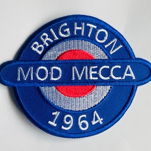 May include: A blue embroidered patch with the words "BRIGHTON MOD MECCA 1964" in white. The patch has a circular design with a red and gray target-like center. The text is in a bold, sans-serif font.