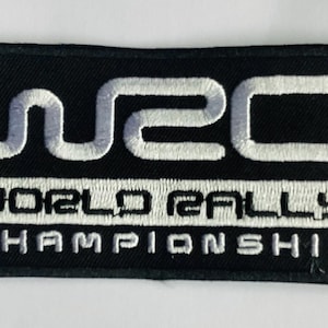Può includere: Toppa rettangolare nera con testo ricamato bianco. Il testo recita "WRC" sopra "WORLD RALLY CHAMPIONSHIP". La toppa ha un bordo nero ed è probabilmente un emblema per le corse di rally.