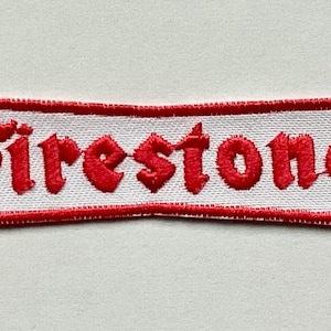 May include: Embroidered patch featuring the word "Firestone" in red, stylized lettering. The text is centered on a white rectangular background with a red border. The patch is likely for clothing or accessories.