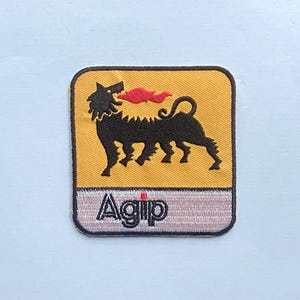 Puede incluir: Parche bordado con el logotipo de Agip. El parche tiene un fondo amarillo con un perro negro de seis patas que respira llamas rojas. La palabra "Agip" está bordada en blanco sobre una pancarta gris rosado.