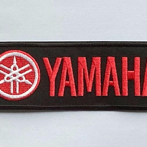 May include: Black rectangular patch with the Yamaha logo and text embroidered in red. The logo is a white emblem inside a red circle, and the word "YAMAHA" is in bold, red letters.