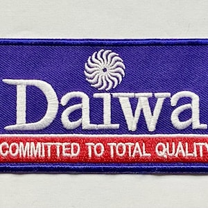 以下が含まれることがあります： 白い「Daiwa」の文字が刺繍されたパッチ。赤いストライプの上に白い文字で「COMMITTED TO TOTAL QUALITY」と書かれています。パッチは青色の背景で、「Daiwa」の文字の上に白い渦巻き模様があります。