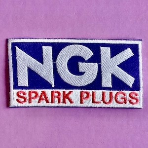 May include: A rectangular embroidered patch featuring the white text NGK on a dark blue background, with the red text SPARK PLUGS positioned below on a white background, all enclosed within a thin white border and stitched fabric edges.