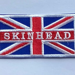 May include: Rectangular embroidered patch displaying the Union Jack flag design. The word "SKINHEAD" is embroidered in white capital letters across the center. The patch has a blue background with red and white details.