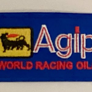 Könnte beinhalten: Ein blaues rechteckiges Abzeichen mit dem Agip-Logo. Das Logo zeigt ein schwarzes, hundeähnliches Wesen in einem gelben Quadrat, mit dem Wort "Agip" in weißer und roter Schrift und "WORLD RACING OILS" in Rot.