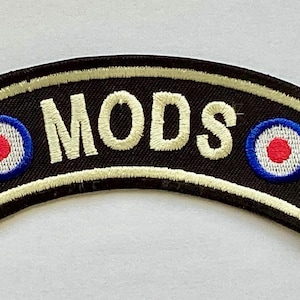 May include: A black embroidered patch with the word "MODS" in white, arched across the top. The patch features two roundels with blue, white, and red concentric circles on each end. The patch has a cream-colored border.