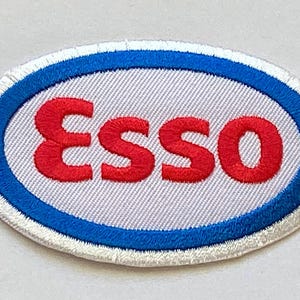 Puede incluir: Parche bordado de forma ovalada con la palabra "Esso" en letras mayúsculas rojas. El texto está centrado sobre un fondo gris claro, enmarcado por un borde azul y blanco. Ideal para ropa o accesorios.