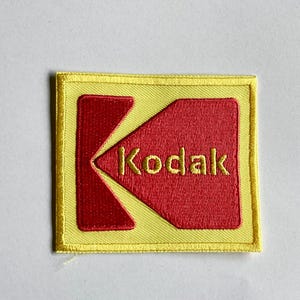 May include: Embroidered patch with the Kodak logo. The patch has a yellow border and a red central design with the word "Kodak" in yellow. The iconic "K" shape is prominent in the design, making it a recognizable emblem.