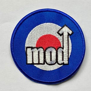 May include: A round, embroidered patch with a blue border. The patch features a white and red target design with the word "mod" in white block letters. An upward-pointing arrow is above the word.