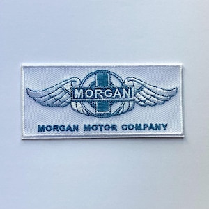 May include: A white rectangular patch with a blue embroidered design. The design includes a circular emblem with the word "MORGAN" and wings. Below the emblem is the text "MORGAN MOTOR COMPANY".