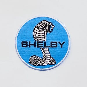 May include: A circular embroidered patch with a coiled cobra design in black and white, set against a light blue backdrop. The word "SHELBY" is embroidered in black block letters beneath the snake.
