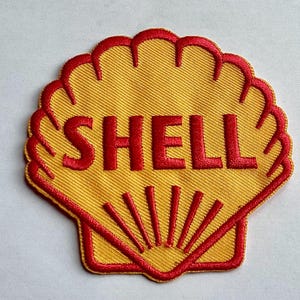 Puede incluir: Parche bordado con el logotipo de Shell. El parche tiene forma de concha de vieira con fondo amarillo y ribete rojo. La palabra "SHELL" está bordada en letras rojas en el centro.