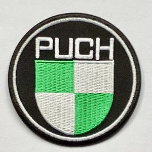 May include: A round, embroidered patch featuring the word "PUCH" in white block letters above a green and white checkered shield design. The patch has a black background with a white border.