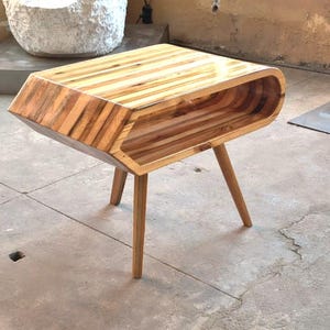 Könnte beinhalten: Ein Beistelltisch aus Holz mit polierter Oberfläche und ausgeprägter horizontaler Maserung. Der Tisch verfügt über ein hohles, oval geformtes Ablagefach und wird von vier konischen Holzbeinen getragen, die nach außen geneigt sind.