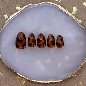 May include: Six tortoise shell-patterned fake nails on a white and gold display stand.