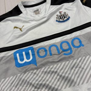 以下が含まれることがあります： 金色のプーマのロゴが入った白と黒のサッカーユニフォーム。ニューカッスル・ユナイテッドの紋章が見えます。シャツには青色の「wonga」の文字と、グレーと白のストライプ模様があります。