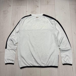 以下が含まれることがあります： 黒いアクセントが付いた白い長袖アスレチックシャツ。 シャツには袖と肩に黒いストライプがあり、袖口と裾にも黒いトリムがあります。 背面の襟には「MUFT」という文字がプリントされています。