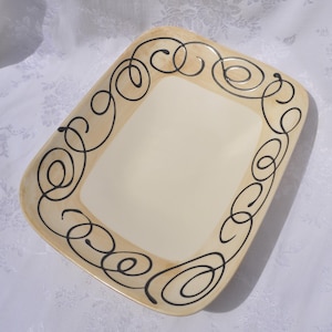 May include: A rectangular ceramic serving platter with a cream-colored center and a wide, light tan border. The border is decorated with a continuous, hand-painted black line pattern consisting of interconnected swirls and loops around the edge.