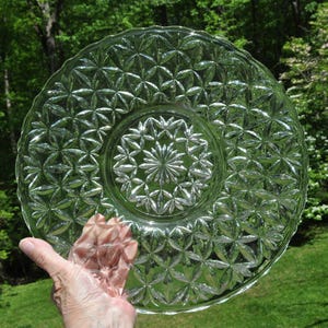 May include: Clear glass serving plate with a starburst pattern in the center and a repeating geometric design. The plate is round with a scalloped edge. The plate is held up in front of a green background.