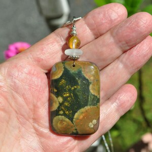 May include: A rectangular pendant made of green, brown, and gold ocean jasper with a white and amber bead accent. The pendant is hanging from a silver chain.
