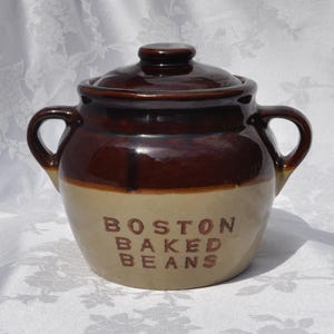 May include: A two-handled ceramic bean pot with a lid, featuring a two-tone design with a dark brown glazed upper section and a tan lower section. The words BOSTON BAKED BEANS are embossed in brown lettering on the front of the tan base.