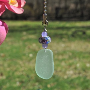 May include: A light green sea glass charm with a purple and white floral bead and a silver chain.