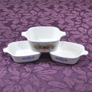 May include: Three white square casserole dishes with handles. One dish features a colorful fruit and vegetable design, while the other two have a blue floral pattern. The dishes are arranged on a purple patterned surface.