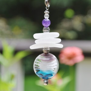May include: A white sea glass pendant with a purple amethyst bead and a silver chain. The pendant is shaped like a sphere and has a layered design with blue and white glass.