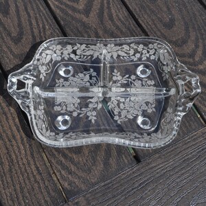 May include: A clear glass serving dish with three compartments and handles. The dish is decorated with a floral pattern. The dish is rectangular with rounded edges and sits on four small feet. The handles are curved and have a decorative design.