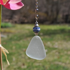 May include: A white sea glass fan pull with a blue and white beaded accent. The fan pull is hanging from a silver chain.