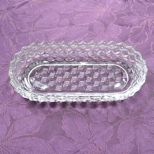 May include: A clear, rectangular glass dish with a diamond pattern on the rim and base. The dish has an oval interior and is likely used for serving or display. The glass is transparent, allowing light to pass through.