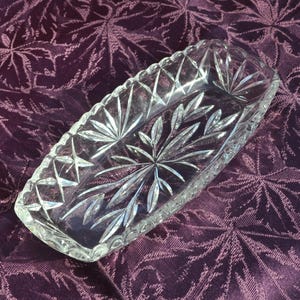 May include: An oval clear glass dish featuring intricate cut patterns. The design includes a central starburst motif surrounded by leaf-like engravings, with a cross-hatched diamond pattern along the scalloped rim and sides of the vessel.