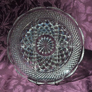 May include: A clear, round glass serving dish with a diamond and starburst pattern. The dish has a scalloped edge and is divided into sections. The dish is displayed on a black metal stand.
