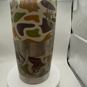 Könnte beinhalten: Edelstahlbecher mit Tarnmuster in Braun-, Grün- und Cremetönen. Der Becher zeigt ein detailliertes geätztes Bild eines Hirsches und das Wort "YETI". Der Becher hat einen transparenten Kunststoffdeckel.