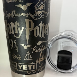 May include: Black stainless steel YETI tumbler with etched Harry Potter-themed designs, including the Sorting Hat, a Golden Snitch, and the words "Harry Potter." The tumbler has a silver rim and a clear lid.