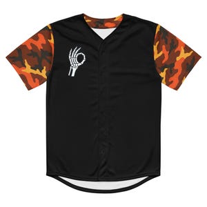 Puede incluir: Camiseta de béisbol negra con mangas de camuflaje naranja, marrón y amarillo. La camiseta presenta una mano de esqueleto blanca haciendo el gesto de "ok" en el pecho izquierdo. La camiseta tiene una parte delantera abotonada y cuello en V.