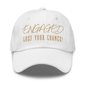 Puede incluir: Gorra de béisbol blanca con las palabras "ENGAGED" y "LOST YOUR CHANCE!" bordadas en hilo dorado. La gorra tiene una visera curva y una correa ajustable en la parte trasera.