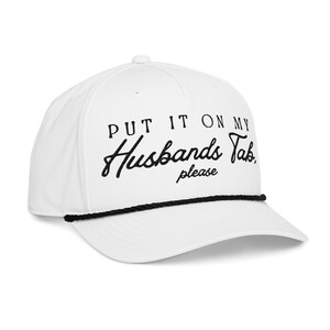 以下が含まれることがあります： 黒いロープのつばと、黒い筆記体で「PUT IT ON MY Husbands Tab, please」と刺繍された白い野球帽。帽子は湾曲したつばと上部にボタンが付いています。