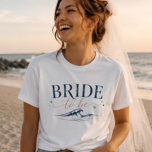Könnte beinhalten: Ein weißes Kurzarm-T-Shirt mit dem dunkelblauen Schriftzug "BRIDE" über dem hellrosa geschriebenen Text "to be". Unter dem Text befindet sich eine Grafik einer dunkelblauen Meereswelle mit kleinen Sternen und einem Herzsymbol.